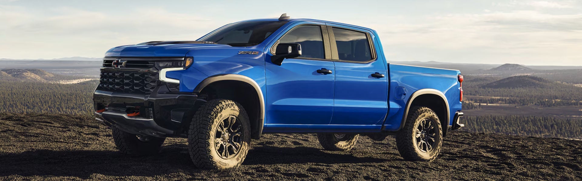 A blue Chevrolet Silverado Zrx pickup truck is parked on a dirt hill, facing slightly left, with a forest and mountains in the background.